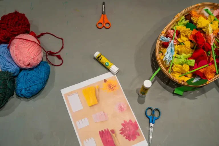 Matériel pour un atelier créatif : pelotes de laine, ciseaux, bâtons de colle et panier rempli de fleurs en papier crépon, accompagnés d'une fiche explicative.