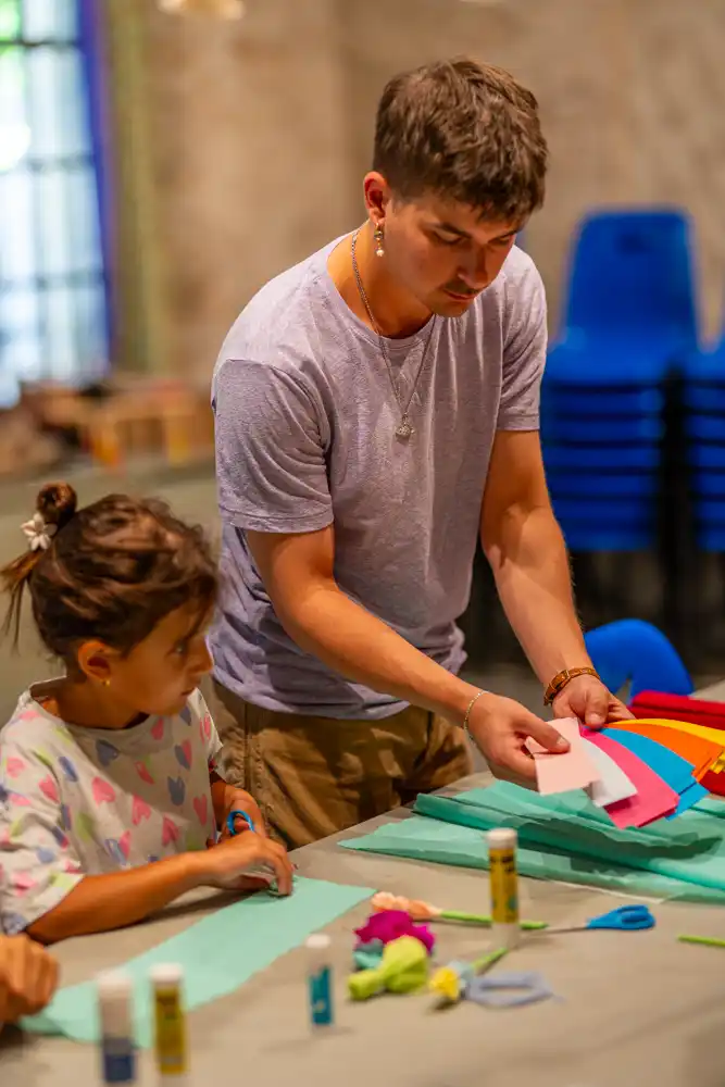 Un animateur aide une enfant à découper du papier coloré lors d'un atelier créatif pour la préparation du corso.