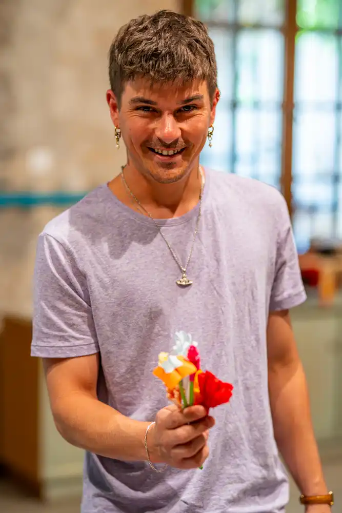 Un jeune homme souriant tenant un petit bouquet de fleurs en papier coloré lors d'un atelier créatif.