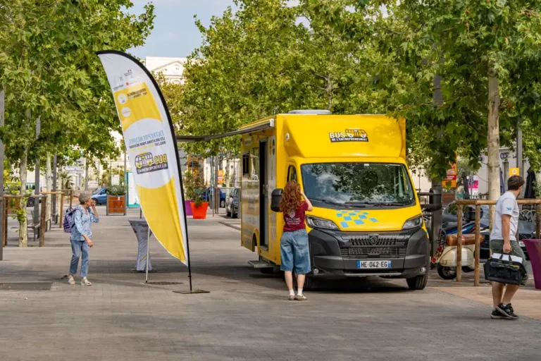 Bus de l'entrepreneuriat stationné sur une place publique avec une oriflamme jaune et blanche à proximité.