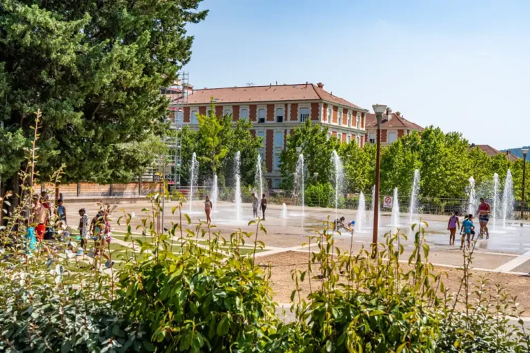 Des enfants et des adultes se rafraîchissent dans les jets d'eau du parc Chabran devant un grand bâtiment en briques.