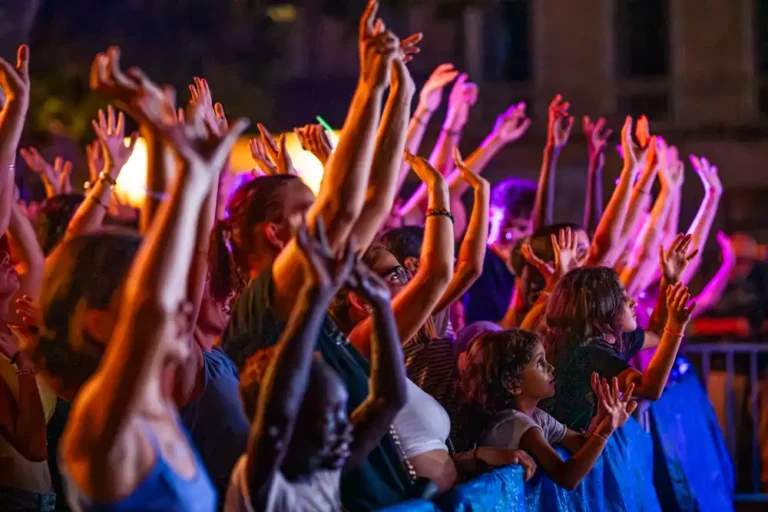 Public nombreux les bras levés lors d'une soirée en extérieur éclairée par des projecteurs colorés.