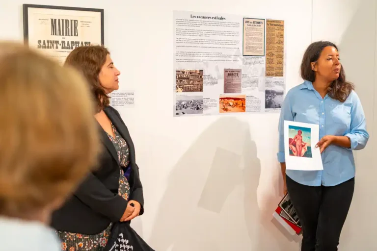 Visite guidée de l'exposition sur l'histoire du tourisme dans le Var aux archives départementales, avec une médiatrice présentant un document illustré devant un panneau explicatif.