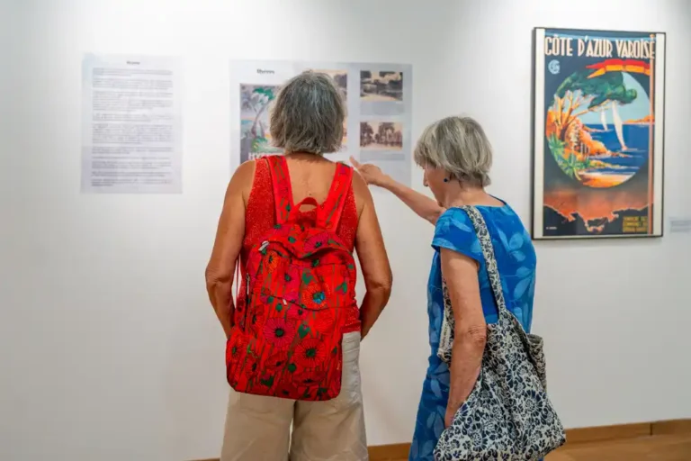 Deux visiteuses observant des panneaux explicatifs et une affiche touristique ancienne lors de l'exposition sur l'histoire du tourisme dans le Var.