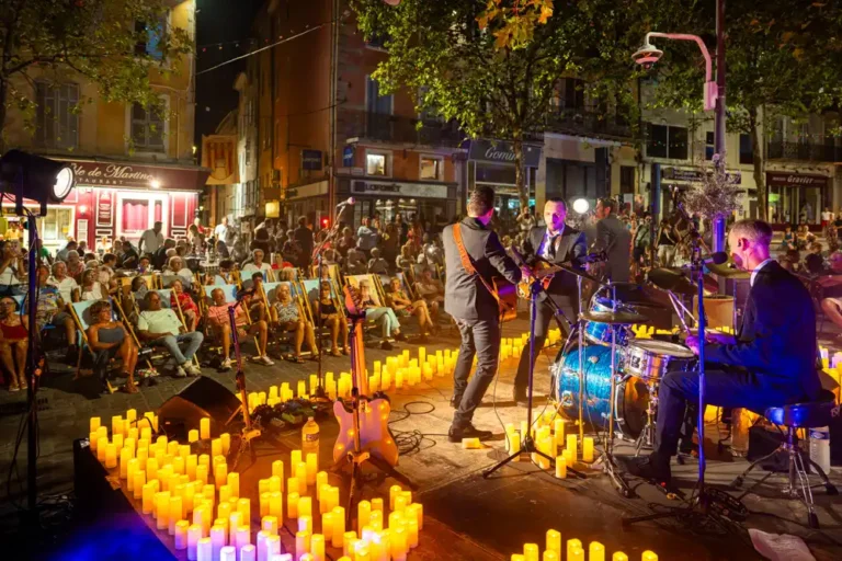 Quatre musiciens en costume jouant sur une scène entourée de bougies devant un public assis sur des chaises longues, place du Marché.