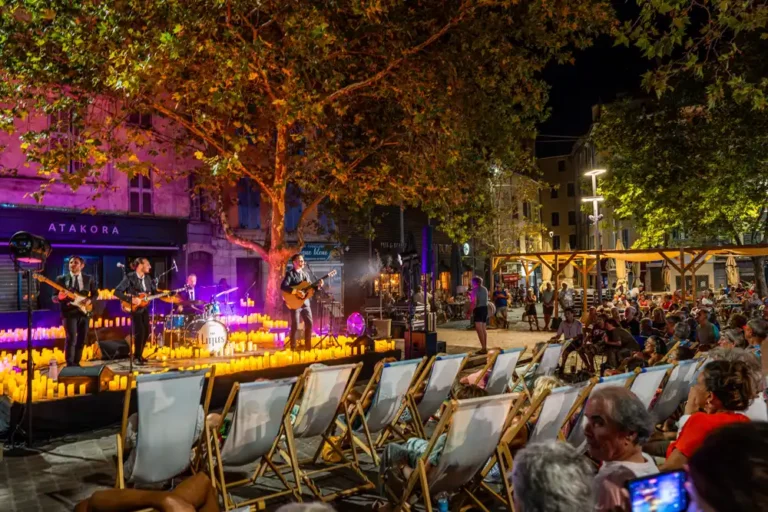 Quatre musiciens en costume sur une scène éclairée par des bougies, devant un public installé sur des chaises longues sur la place du Marché.