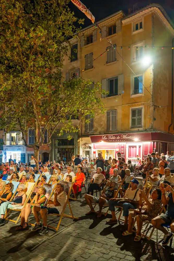 Public assis sur des chaises longues sur la place du Marché lors d'un concert en soirée.