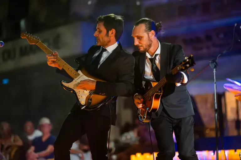Deux musiciens en costume jouant de la guitare électrique et de la basse lors d'un concert en extérieur sur la place du Marché.