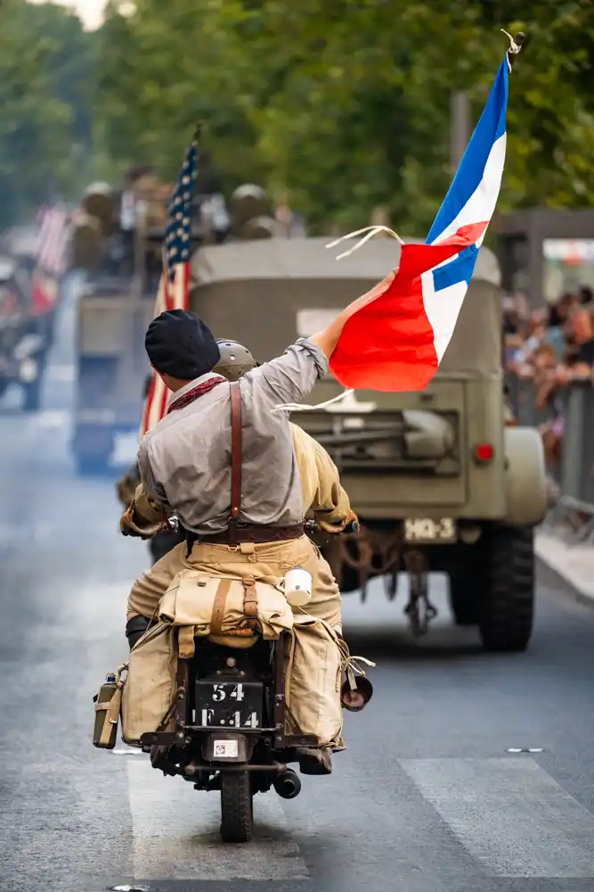 Participant en tenue d'époque sur une moto militaire, agitant un drapeau français lors du défilé de la Libération sur le boulevard Clemenceau.