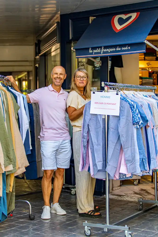 Un commerçant et une commerçante souriants devant leur boutique « Le petit français du sud » lors de la braderie, à côté d'un portant de vêtements pour hommes affichant une réduction de 50 %.