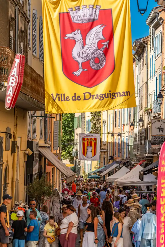 Rue commerçante animée de Draguignan lors de la braderie, avec des stands sous des tentes et des drapeaux aux couleurs de la ville suspendus au-dessus de la foule.