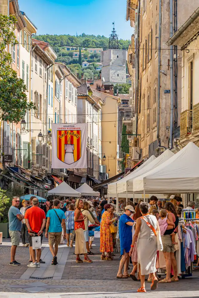 Foule de visiteurs déambulant dans une rue commerçante piétonne de Draguignan lors de la braderie, avec des stands sous chapiteaux blancs et une bannière du quartier historique Lei Fadoli.