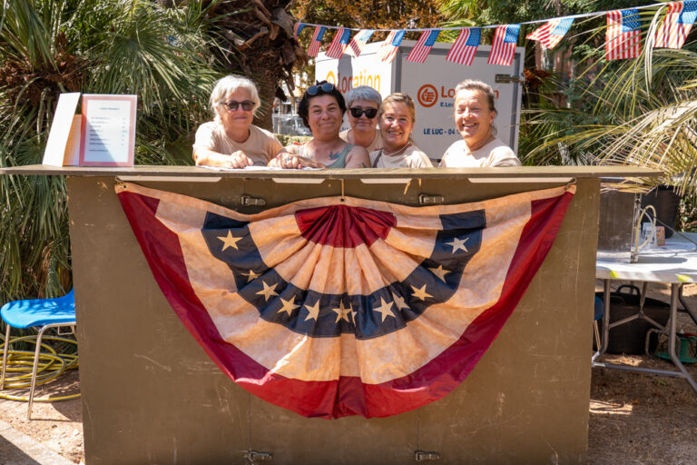 Cinq femmes souriantes derrière un comptoir décoré d'une guirlande aux couleurs des États-Unis, lors de la reconstitution d'un camp militaire au jardin Anglès.
