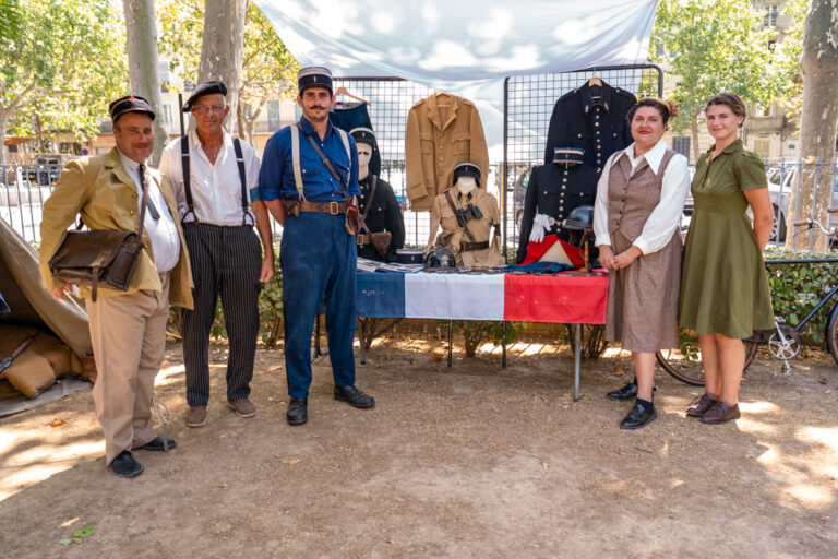 Reconstitution historique au jardin Anglès : cinq personnes en tenue d'époque présentent des uniformes et accessoires militaires sur un stand.