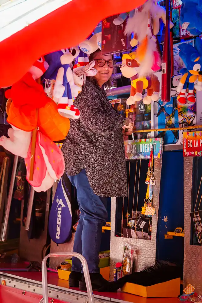 Une femme souriante derrière le comptoir d'un stand de fête foraine rempli de peluches et de lots à gagner.