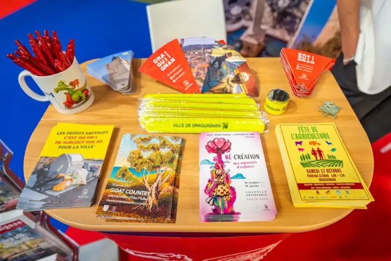 Table d'information lors du salon des maires présentant des brochures municipales, des dépliants d'expositions culturelles, des bracelets réfléchissants et des objets promotionnels de la ville de Draguignan.