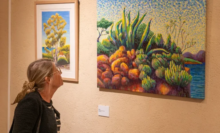 Une visiteuse observe une peinture colorée représentant un paysage côtier avec des agaves, exposée à la Chapelle de l’Observance.