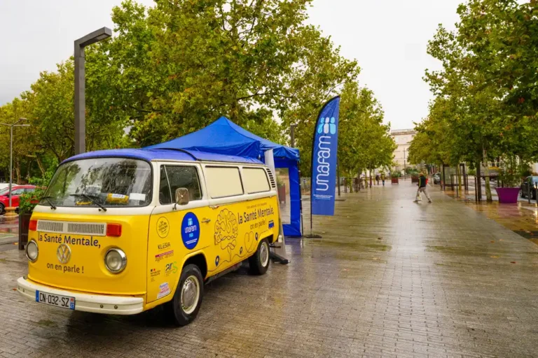 Combi Volkswagen jaune stationné sur une allée pavée, accompagné d'un barnum bleu et d'une oriflamme Unafam pour une opération de sensibilisation à la santé mentale.