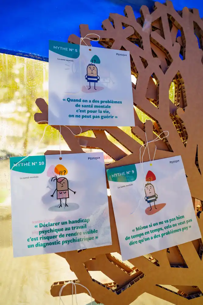 Trois panneaux d'information suspendus sur un support en carton, présentant des mythes sur la santé mentale illustrés par Psycom : 'Quand on a des problèmes de santé mentale c'est pour la vie', 'Déclarer un handicap psychique au travail c'est risquer de rendre visible un diagnostic psychiatrique' et 'Même si on ne va pas bien de temps en temps, cela ne veut pas dire qu'on a des problèmes psy'.